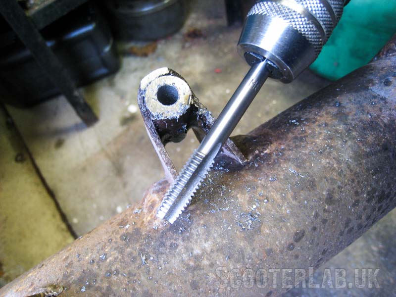 Create a thread in the hole with an M8 tap. If the tap is tapered then you may need to tap deeper than you expect for the pin to wind all the way in without resistance. You need a short-handled tap-wrench to be able to fully rotate the tap without touching the legshield support strut. Lubricate the tap and turn the tap back by a quarter turn for every half turn you go forward.